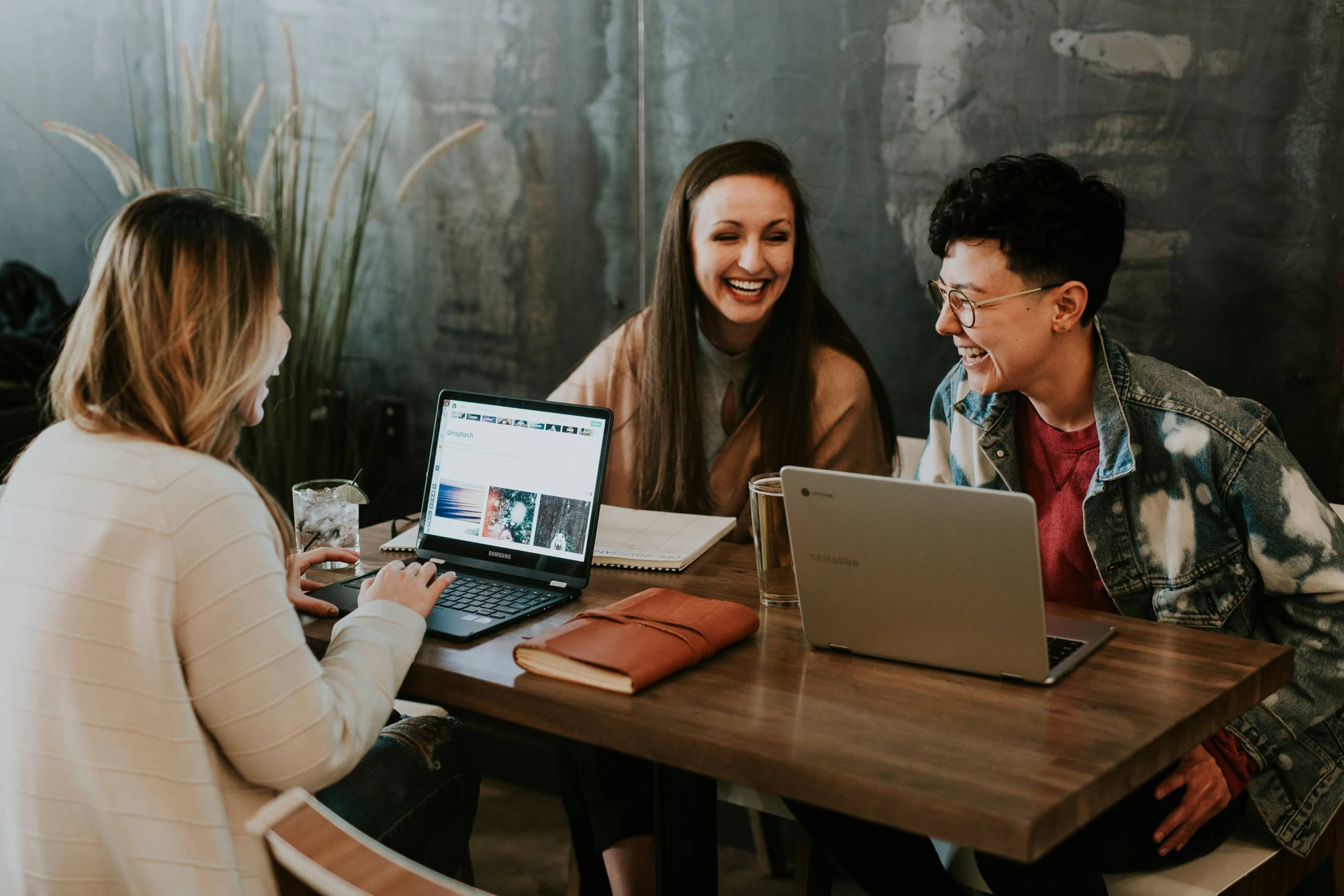 Smiling young women using laptops in a modern coffee shop for teamwork and customer experience discussion.